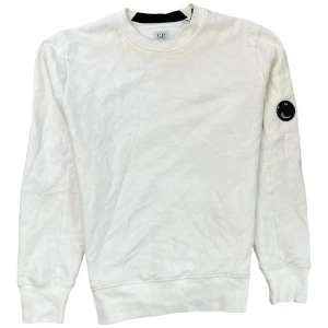 Vit sweatshirt från C.P. Company - Säljer en stilren vit sweatshirt från C.P. Company. Tröjan har en rund halsringning och en cool detalj med en logotyp på ärmen. Perfekt för en avslappnad stil.