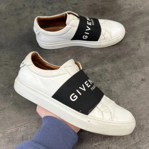 Vita sneakers från Givenchy - Snygga vita sneakers från Givenchy med en bred svart rem över foten där märkesnamnet står i vitt. Skorna har en stilren design med rund tå och är tillverkade i skinn. Perfekta för en trendig look.