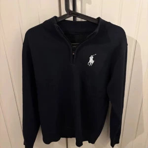 Mörkblå tröja med dragkedja från Ralph Lauren - Säljer en stilren mörkblå tröja från Ralph Lauren med en liten dragkedja vid halsen. Tröjan har en broderad logotyp på bröstet och långa ärmar. Perfekt för en avslappnad men snygg look.