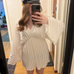 Studentklänning  - Säljer en vit klänning med plisserade detaljer och v-ringning. Klänningen har långa, vida ärmar. Perfekt för student eller liknande evenemang. Aldrig använd med lapp kvar på