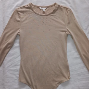 Beige långärmad topp med strassdetaljer - Säljer en elegant beige långärmad topp med strassdetaljer vid halsringningen. Perfekt för att lägga till lite glans till din outfit. Toppen har en rund hals och är tillverkad i ett mjukt material.