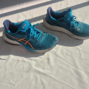 Blå löparskor från Asics - Säljer ett par blå löparskor från Asics med orange detaljer. Skorna har snörning och en bekväm passform, perfekt för träning. De är tillverkade i syntetmaterial och har en rund tå.