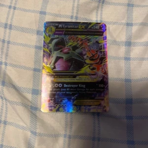 Pokémonkort Mega Tyranitar EX - Säljer ett Pokémonkort av Mega Tyranitar EX med 240 HP. Kortet har en holografisk yta med färgglada detaljer och visar en kraftfull Tyranitar i strid. Perfekt för samlare eller spelare som vill förstärka sin kortlek.
