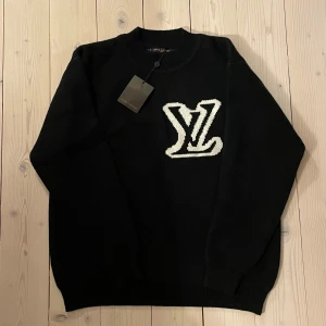 Svart tröja från Louis Vuitton - Har nu fått in en ny snygg svart tröja från Louis Vuitton med ett stort vitt LV-märke på bröstet. Tröjan har långa ärmar och en klassisk rund halsringning. Perfekt för en stilren look. Storlek S men passar som XL