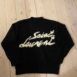 Svart tröja från Saint Laurent - har fått in en ny snygg, svart tröja från Saint Laurent med ett snyggt vitt mönster på framsidan. Tröjan har långa ärmar och en klassisk rund halsringning. Perfekt för en stilren look. Tröjan är i storlek S men passar L