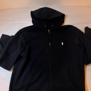 Svart hoodie från Polo Ralph Lauren - Säljer en stilren svart hoodie från Polo Ralph Lauren med dragkedja och det klassiska broderade logotypen på bröstet. Perfekt för en avslappnad look. Den har långa ärmar och en praktisk huva.