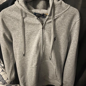Grå hoodie från Ralph Lauren - Säljer en stilren grå hoodie från Ralph Lauren med dragkedja och klassisk logga på bröstet. Perfekt för en avslappnad look. Den har en justerbar huva och fickor framtill.