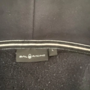 Svart hoodie från Sail Racing - Säljer en svart hoodie från Sail Racing med dragkedja och huva. Jackan har en ficka med dragkedja på bröstet och är långärmad. Perfekt för en avslappnad stil.