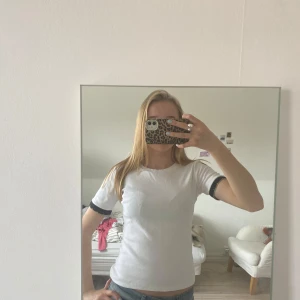 Vit t-shirt med svarta detaljer - Säljer en stilren vit t-shirt med svarta kanter på ärmarna. Perfekt för en casual look. Passar bra till jeans eller shorts. Hör av dig vid intresse!