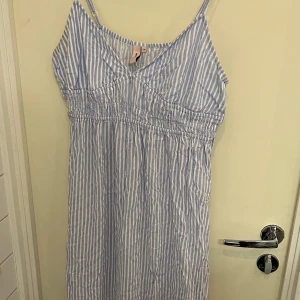 Blå- och vitrandig klänning - Söt klänning med smala axelband och v-ringning. Den har ett randigt mönster i blått och vitt och en smockdetalj under bysten för en bekväm passform. Perfekt för varma dagar.