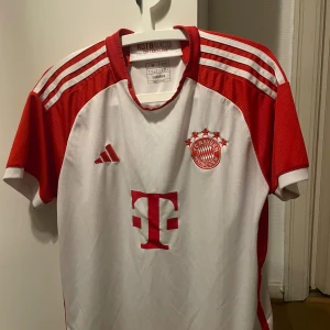 Bayern München tröja - Säljer en vit och röd fotbollströja från Adidas med FC Bayern München-logga och spelarnamn på baksidan. Tröjan har korta ärmar och klassiska ränder på axlarna. Perfekt för fans av laget!