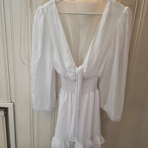 Vit klänning med volangdetaljer - Säljer en söt vit klänning med v-ringning och volangdetaljer. Klänningen har långa ärmar och ett knytband i midjan för en fin passform. Perfekt för sommaren