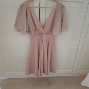 Champagnefärgad klänning från Goddiva - Säljer en elegant champagnefärgad klänning från Goddiva med v-ringning och korta, fladdriga ärmar. Klänningen har en fin passform och är perfekt för speciella tillfällen. Den har en lätt glittrig yta som ger en extra touch av glamour.