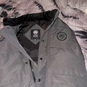 Grå dunväst från Canada Goose - Säljer en stilren grå dunväst från Canada Goose med svart krage och dragkedja. Västen har en broderad logotyp på bröstet och knappar framtill. Perfekt för kyliga dagar.