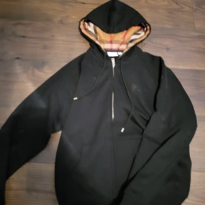 Svart hoodie från Burberry - Säljer en stilren svart hoodie från Burberry med dragkedja och den klassiska Burberry-rutiga fodret i huvan. Perfekt för en avslappnad och trendig look.
