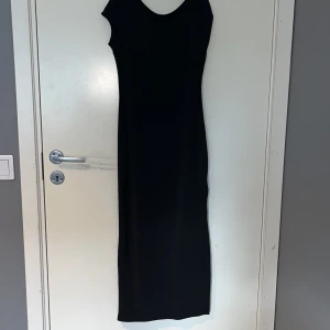 Svart långklänning med slits - En stilren svart långklänning med rundad halsringning och ärmlös design. Klänningen har en snygg slits på sidan och en öppen rygg, vilket ger en elegant och modern look. Perfekt för dig som gillar minimalistisk stil.