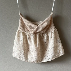 Beige broderad linnetopp med spets - Supersöt beige linnetopp med broderade blommor och spetsdetaljer. Toppen har en omlottliknande överdel i ribbat tyg. Perfekt för dig som gillar romantisk och bohemisk stil!