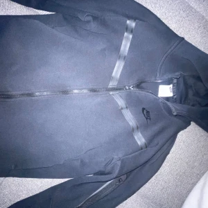 Svart Nike vindjacka med dragkedja L - Svart Nike tech fleece i storlek 147-158cm. Den  har hel dragkedja framtill, huva och diskreta svarta detaljer över bröstet. Tillverkad i ett mjukt syntetmaterial, perfekt för chill eller när du är på språng. Klassisk Nike-logga på bröstet.