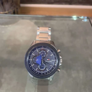 Tommy Hilfiger silverfärgad klocka - Stilren klocka från Tommy Hilfiger med silverfärgad länk i metall och mörkblå urtavla. Klockan har flera små urtavlor för dag och datum, samt tydliga vita markeringar och logga. Perfekt accessoar för dig som gillar klassisk design med sportig touch.