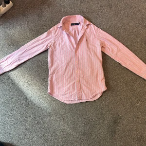 Rosa randig skjorta från Ralph Lauren - Säljer en klassisk rosa skjorta från Ralph Lauren med vita ränder och broderad logga på bröstet. Skjortan har lång ärm, knappar framtill och en snygg krage. Perfekt för dig som vill elvolvera sin stil.