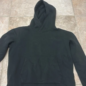 Svart hoodie från Sail Racing - Svart hoodie från Sail Racing med klassisk känguruficka och broderad logga på bröstet. Tröjan har huva och är tillverkad i mjuk bomullsmix, perfekt för en chill och avslappnad stil. Långärmad modell med ribbade muddar vid ärmslut och nederkant.