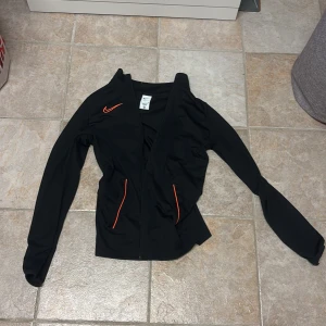 Svart Nike vindjacka med orange detaljer - Svart vindjacka från Nike med orange swoosh på bröstet och orange detaljer vid fickorna. Jackan har dragkedja framtill och är tillverkad i lätt polyester, perfekt för aktiva dagar. Snygg och sportig look med lång ärm och smal passform.