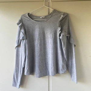 Grå långärmad tröja med volang - Säljer en ljusgrå långärmad tröja med volangdetaljer längs axlarna och ärmarna. Tröjan har en rund halsringning och är tillverkad i mjukt bomullsmaterial. Perfekt för dig som gillar en enkel men ändå trendig look.