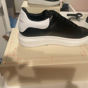 Alexander McQueen svarta sneakers - Stilrena svarta sneakers från Alexander McQueen med vit häl och tjock vit sula. Skorna har snörning, rund tå och är tillverkade i skinn. Perfekta för dig som gillar en clean och lyxig look med tydlig logga på hälen.