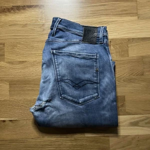 Replay Anbass hyperflex jeans - Säljer ett par blå Replay jeans med klassisk femficksdesign och snygga slitningar. Jeansen har raka ben och tydliga slitdetaljer på baksidan. Mörkblå tvätt med ljusare partier och diskret logga på bakfickan. Perfekta för en avslappnad streetstil. Längd 106cm midja 41cm! Defekt i grenen som syns tydligare på bild 4