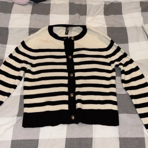 Randig kofta från H&M Divided - Säljer en svartvit randig kofta från H&M Divided i storlek S. Koftan är ribbstickad med guldiga knappar framtill och har långärmad passform. Perfekt för lager-på-lager och ger en klassisk vibe till din outfit.