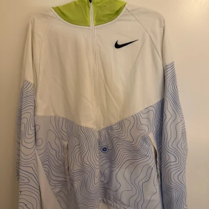Vit och limegrön Nike vindjacka - Nike vindjacka i vitt med limegrön krage och blått vågigt mönster på ärmar och nedre delen. Jackan har hel dragkedja framtill och svart Nike-logga på bröstet. Perfekt för dig som gillar sportig och unik stil.