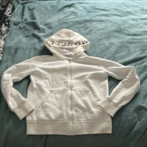 Vit Moncler hoodie med dragkedja - Äkta Från moncler.Snygg vit hoodie från Moncler med dragkedja framtill, stor huva med Moncler-tryck och klassisk logga på ärmen. Tröjan har kängurufickor och ribbade muddar. Perfekt för dig som gillar stilrena och exklusiva plagg.
