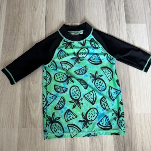 Grön badtröja med fruktmönster H&M - Cool badtröja från H&M i storlek 122/128. Framsidan är grön och blå med svarta ananas- och vattenmelonmönster, medan ärmarna är svarta med gröna sömmar. Perfekt för strand eller poolhäng i sommar!