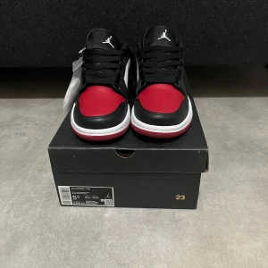 Nike Air Jordan 1 Low röd/svart/vit - Nike Air Jordan 1 Low i klassisk röd, svart och vit färg. Skorna har svart snörning, vit mellansula och röd yttersula.  Tillverkade i skinn och syntet, med rund tå och platt sula. Extra vita skosnören medföljer. Aldrig använda i storlek 38, säljs pga för stora. Hör av er vid minsta fundering.