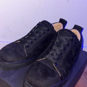 Svarta mockasneakers från Louboutin - Snygga svarta sneakers i mocka från Christian Louboutin med ikonisk röd sula och broderad logga bak. Skorna har svarta snören, rund tå och beige innersula med guldfärgad logotyp. Perfekta för dig som vill sticka ut med exklusiv streetstyle.