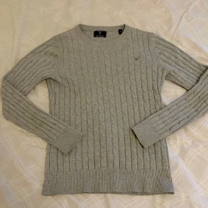 Grå kabelstickad tröja från Gant - Snygg grå kabelstickad tröja från Gant med rund hals och diskret logga på bröstet. Tröjan har lång ärm och ribbade muddar, perfekt för lager på lager under kyliga dagar. Klassisk och stilren modell som funkar till allt.