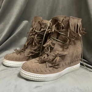 Fransskor - Unika sneakers i beige mocka med massor av fransdetaljer och snörning framtill. Skorna har en vit platt sula med mönstrad kant och grova stickningar längs kanterna. Perfekta för dig som vill sticka ut med en boho-inspirerad stil.