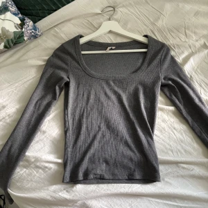 Grå ribbad topp från Nelly - Snygg grå långärmad topp från Nelly med ribbad struktur och rundad, djup urringning. Perfekt att styla med jeans eller kjol för en clean look. Mjuk och stretchig, sitter tajt mot kroppen och är enkel att matcha.