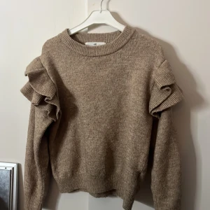Beige/brun stickad volangtröja från H&M - Superfin beige stickad tröja från H&M med volangdetaljer på axlarna. Tröjan har rund halsringning, ribbade muddar och lång ärm. PRIS KAN DISKUTERAS 