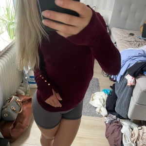 Vinröd stickad tröja med guldknappar - Vinröd stickad tröja med lång ärm och snygga guldknappar vid ärmslutet. Tröjan har en klassisk rund halsringning och är i en mjuk, skön kvalitet som passar perfekt till höst och vinter. Enkel att matcha med både jeans och kjol. Kommer från ZARA och är i storlek S.