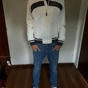 Vit vindjacka från Sail Racing - Vit vindjacka från Sail Racing med marinblå och röda detaljer. Jackan har dragkedja framtill, två fickor med dragkedja och ribbade muddar i ärmslut och nederkant. Snygg sportig look med broderad logga på bröstet. Perfekt för dig som gillar stilren och funktionell design.