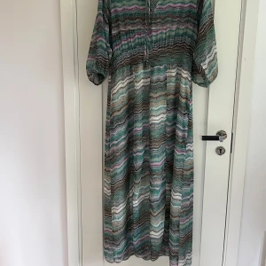 Mönstrad långklänning Line of Oslo - Säljer en långklänning från Line of Oslo med vågigt mönster i grönt, brunt, vitt och lila. Klänningen har trekvartsärmar, v-ringning med knyt och luftig passform.  Fastsydd underkjol. Perfekt för dig som gillar bohemisk stil och färgkombinationer.