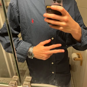 Ralph lauren skjorta  -  Strlk S-M Klassisk mörkblå skjorta från Polo Ralph Lauren i 100% bomull. Skjortan har långa ärmar, knappar framtill och ikonisk röd broderad logga på bröstet. Perfekt för dig som gillar stilrena och tidlösa plagg med snygga detaljer.