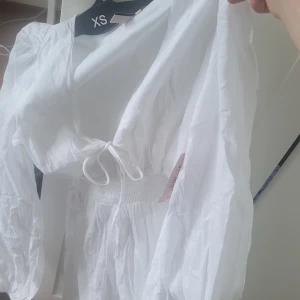 Vit kortklänning från Nelly med knyt - Superfin vit kortklänning från Nelly i luftig bomull. Klänningen har långa ärmar, smockad midja och knyt framtill. Perfekt för varma dagar med sin lätta känsla och snygga passform. Enkel men ändå trendig design som passar till många tillfällen.