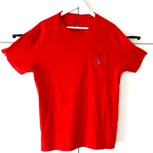 Polo Ralph Lauren porsche röd t-shirt, storlek M-L - Röd t-shirt från Polo Ralph Lauren med klassisk passform. T-shirten har rund hals, korta ärmar och en bröstficka med blå broderad logga. Tillverkad i mjuk bomull som känns skön mot huden. Perfekt för en clean och stilren look.Mycket fin skick!             Ordpris 1000kr.