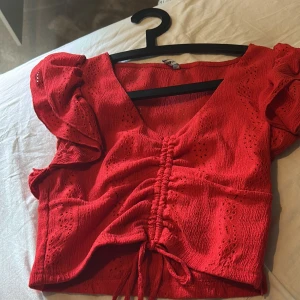 Röd croppad blus med volangärm - Superfin röd croppad blus från Primark med v-ringning, volangärmar och snygg snörning framtill. Blusen har broderade detaljer och är tillverkad i ett lätt, luftigt tyg som passar perfekt till sommaren. Perfekt för dig som vill sticka ut med färg och coola detaljer!