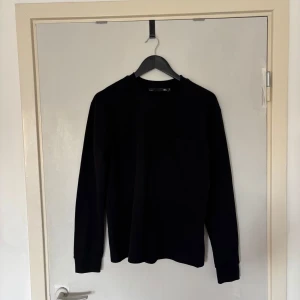 Svart långärmad tröja 157 S - Enkel svart långärmad tröja från 157 i storlek S. Tröjan har rund halsringning och en bröstficka som ger en clean och stilren look. Perfekt till jeans eller cargo pants. Materialet är mjukt och känns skönt mot huden.
