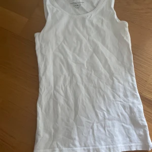 Vit linnetopp från Newbody XS - Klassisk vit linnetopp från Newbody i storlek XS. Toppen är ärmlös med rund halsringning och har en slim passform. Tillverkad i mjuk och stretchig bomull med 5% elastan för extra komfort. Perfekt att styla till jeans eller shorts.