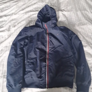 Marinblå vindjacka Tommy Hilfiger - Snygg marinblå vindjacka från Tommy Hilfiger med huva och hel dragkedja med rödvit detalj. Jackan har ribbade muddar, två fickor fram och diskret logga på bröstet. Perfekt för blåsiga dagar och enkel att matcha med det mesta.