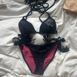 Svart och rosa bikini från Odd Molly - Snygg bikini från Odd Molly i svart med rosa foder och broderade detaljer. Bikinin har knytband i flätad modell både upptill och i sidorna på underdelen. 